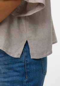 Chemise en lin gris clair avec un ourlet arrondi, des fentes sur les côtés et une coupe décontractée, associée à un jean en denim bleu foncé. Détails de couture visibles.