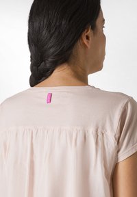 Ljusrosa blus med en sammanknuten ryggdesign, korta ärmar och en liten lila etikett vid halsringningen. Slät textur.