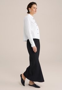 Camisa blanca de corte corto con botones negros, combinada con una falda negra ajustada. Llevada con zapatos negros sin cordones. Textura de tela suave.