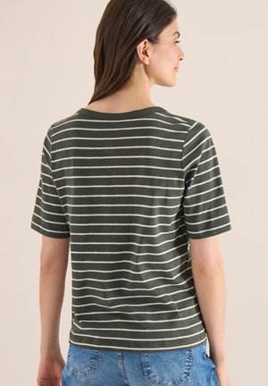Femme aux longs cheveux bruns portant un t-shirt à manches courtes rayé vert foncé et blanc, ainsi qu'un jean bleu, face à droite sur un fond neutre.