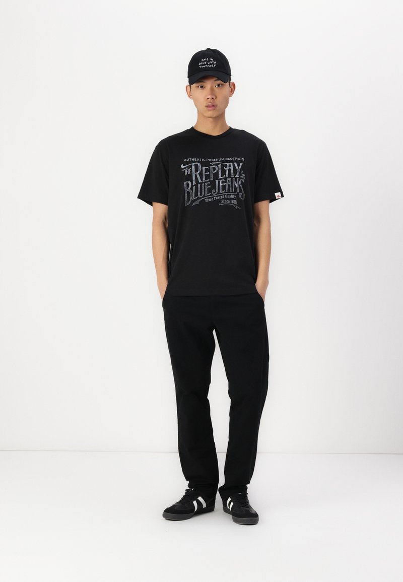 Schwarzes T-Shirt mit weißem Textdesign, kombiniert mit schwarzen Hosen und schwarzen Sneakers mit weißen Streifen. Die Accessoires umfassen eine schwarze Mütze.