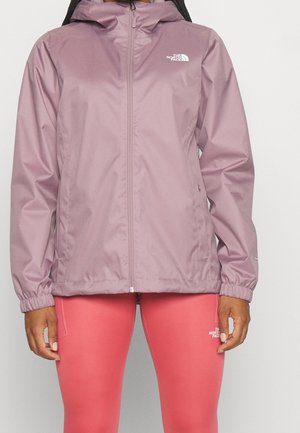 Kobieta ubrana w jasnofioletową kurtkę zapinaną na zamek oraz koralowe legginsy, na obu widoczne loga The North Face na klatce piersiowej i udzie.
