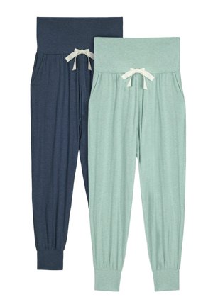 Zwangere vrouw staat op blote voeten, draagt een wit mouwloos topje en lichtgroene zwangerschapsjoggingbroek met zijzakken tegen een effen achtergrond.