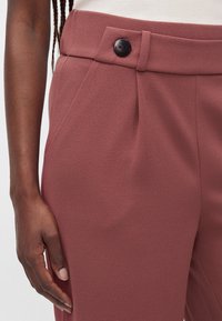 Gros plan sur la main d’une personne reposant à ses côtés, vêtue d’un pantalon taille haute mauve avec un bouton noir sur la ceinture.