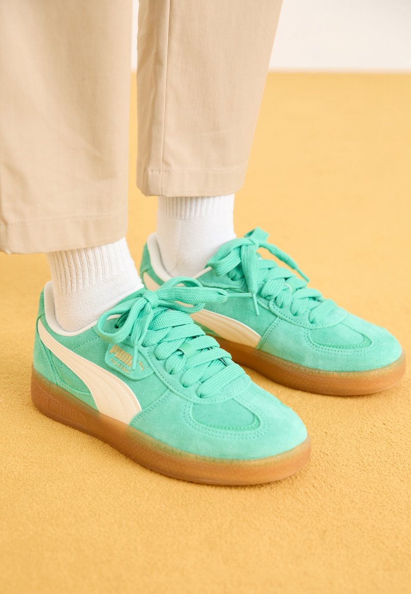 Baskets en daim turquoise avec des accents blancs, bout rond et lacets assortis. Semelle en caoutchouc texturée de couleur naturelle. Portées avec un pantalon beige.