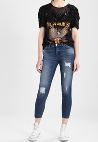 Mujer que lleva una camiseta gráfica de Milwaukee negra con adornos, jeans ajustados azules desgastados y zapatillas altas blancas.