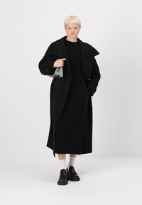 Casaco de lã preto com um colarinho largo, combinado com um vestido de malha preto. A modelo usa sapatos pretos e meias brancas, segurando uma pequena bolsa.