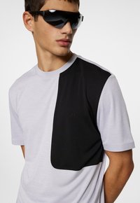 Grå och svart kortärmad t-shirt med en slät yta, en stor svart lapp på vänster sida och rund halsringning.