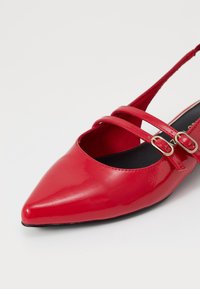 Scarpa in pelle verniciata rossa con punta affusolata, dotata di due cinturini incrociati e dettagli con fibbie dorate. Rivestimento interno e suola nera.