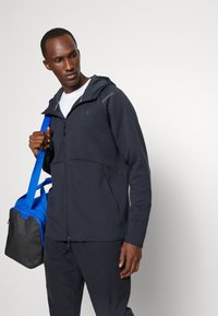 Giacca a zip da uomo grigio scuro con cappuccio, caratterizzata da una texture liscia e un logo tonale, abbinata a una borsa a tracolla blu e nera.