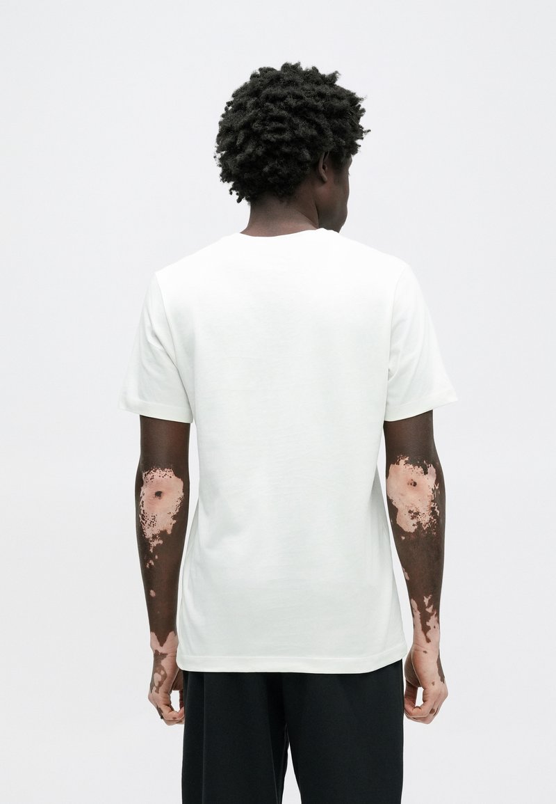 Homme à la peau foncée et au vitiligo sur les bras porte une chemise blanche à manches courtes unie et un pantalon noir, debout de dos sur un fond neutre.