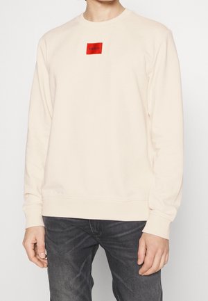 Person trägt einen cremefarbenen Pullover mit einem kleinen roten quadratischen Logo mit der Aufschrift "HUGO" auf der Brust und dunkelgraue Jeans.