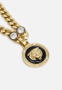 Gouden ketting met een zwarte ronde hanger met een verheven gouden tijderkop en kristalaccenten; branding gegraveerd rond de rand van de hanger.
