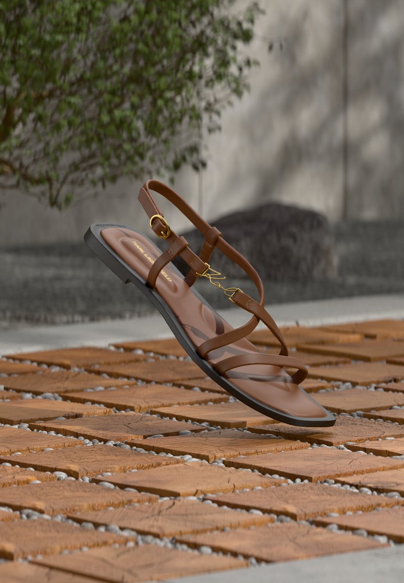 Καφέ επίπεδη σαγιονάρα με λουράκια και λεπτομέρεια με χρυσή αγκράφα, αιωρείται πάνω από ξύλινο πλακάκι σε εξωτερικό χώρο με φυτά στο παρασκήνιο.