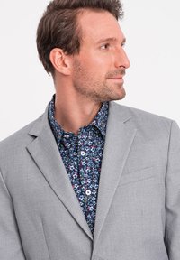 Vaaleanharmaa bleiseri tummansinisellä kukkapaidalla, jossa on valkoiset napit. Bleiserissä on yksi rintatasku ja muotoon ompeloitujen kaulusten yksityiskohta.