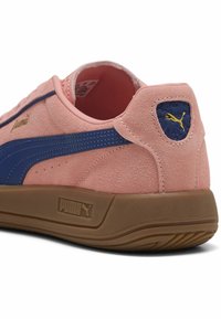 Zapatilla de ante rosa con acentos azules, que presenta un logo dorado y un parche texturizado en el talón. Suela de goma y forro suave.