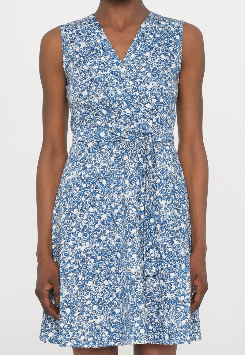 Robe sans manches bleue et blanche à fleurs, à encolure en V et ceinture à la taille, portée par une personne sur un fond uni.