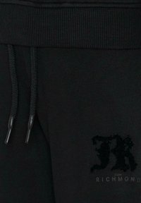 Zbliżenie na pas czarnych dresów z sznurkiem i puszystym haftowanym logo "JR" nad napisem "John Richmond" na tkaninie.