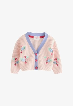 Junges Mädchen in einem bestickten rosa Cardigan, gestreiften weiten Hosen und roten Schuhen steht drinnen neben Pappblumen und gestreiftem Vordach.