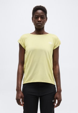 Stojaca osoba s krátkymi kučeravými vlasmi oblečená v svetložltej krátkemu rukávu a čiernych nohaviciach na jednoliatom pozadí.