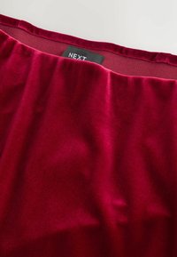 Tela de terciopelo rojo con una textura suave, que presenta una cinturilla con fruncidos. Etiqueta de la marca "NEXT" visible cerca del borde superior.