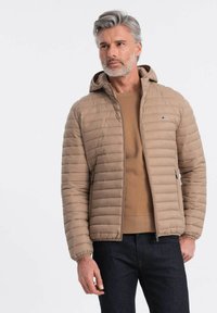 Beige polstret jakke med hætte, der har lodret quiltning, frontlynlås, sidelommer og et lille logo på brystet. Båret over en tan sweater.