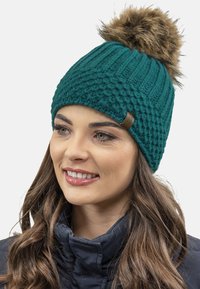 Gorro de punto teal con patrón texturizado, que presenta un pompón de peluche marrón en la parte superior y una pequeña etiqueta de cuero en el lateral.