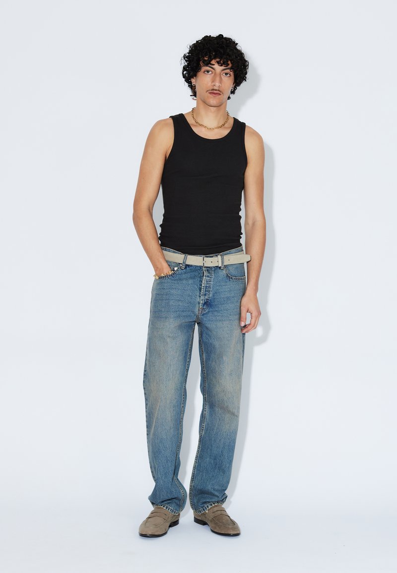 Junger Mann mit lockigen Haaren, der ein schwarzes Tanktop, hellblaue Jeans, einen beigen Gürtel, beigen Schuhe und goldene Kettenschmuck trägt.