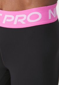 Černé legíny s širokým růžovým pasem, na kterém je bílým písmem nápis "NIKE PRO". Hladká textura a přiléhavý střih.