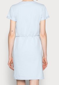 Kobieta w jasnoniebieskiej sukience z krótkim rękawem i paskiem w talii, pokazana od tyłu na jednolitym tle.