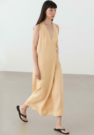 Femme marchant dans une robe midi jaune pâle sans manches à col halter avec des tongs noires sur un sol neutre et un fond blanc.