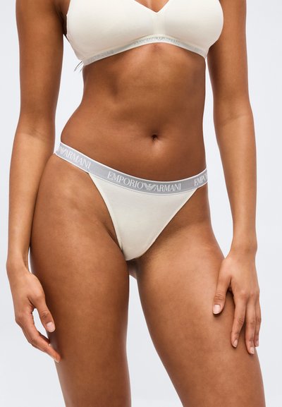 Γυναίκα που φοράει λευκή εσώρουχα Emporio Armani, εμφανίζοντας τον κορμό και τα επάνω μέρη των ποδιών σε απλό φόντο.