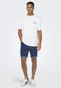 T-shirt bianco in cotone con grafica verde sul petto, abbinato a pantaloni shorts in denim blu con risvolti e sneakers bianche.