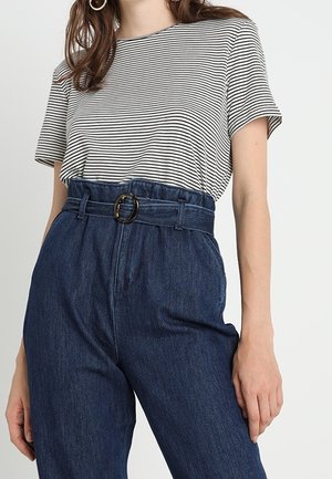 Nainen, joka pukeutunut tummansinisiin korkeavyötäröisiin farkkuihin, joissa on vyö, ja mustavalkoraidalliseen lyhythihaiseen paitaan, seisomassa yksiväristä taustaa vasten.