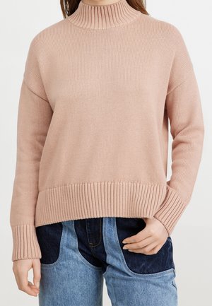 Pull à mailles rose clair avec un col montant côtelé, des épaules tombantes et un ourlet côtelé, associé à un jean en denim bleu.
