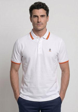 Hombre con camiseta polo blanca con ribete rojo y amarillo en el cuello y las mangas, de pie con las manos en los bolsillos de pantalones azul marino.
