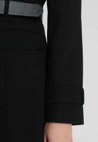 Gros plan d'une manche de manteau noire avec un détail de lanière au niveau du poignet et une partie d'une ceinture noire à la taille.