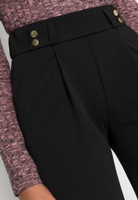 Close-up van zwarte high-waist broek met gouden knopen, gedragen met een textuur van een lila langemouwen top, waarbij de taille en een deel van de arm zichtbaar zijn.