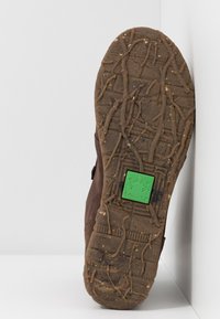 Ruskea kenkäpohja, jossa on kuvioitu kumimateriaali ja vihreä sammakko-logo. Muotoilussa on uria, jotka parantavat pitoa ja kestävyyttä.