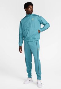 Τιρκουάζ φόρμα από λείο ύφασμα, που περιλαμβάνει μπουφάν με φερμουάρ και όρθιο γιακά, καθώς και παντελόνι με αντίθετες πλευρικές ρίγες.