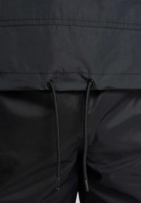 Czarna kurtka przeciwwiatrowa o gładkiej fakturze, wyposażona w regulowany ściągacz w talii oraz bezszwowy design na dole.