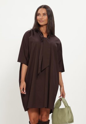 Woman wearing loose dark brown dress with half sleeves and a tied neckline, holding a light green handbag, standing against white background.