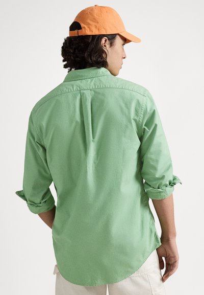Ljusgrön långärmad skjorta med uppvikta ärmar; mjuk textur. Orange keps. Sedd bakifrån, med detaljer som krage och knappar.