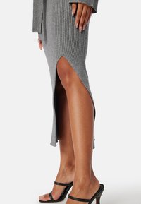 Šedé žebrované pletené šaty s vysokým bočním rozparkem, s úzkým střihem a dlouhými rukávy. Nosené s černými sandály na suchý zip.