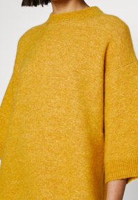 Femme portant un pull en maille pelucheux jaune moutarde, aux manches trois-quarts et col rond, sur un fond uni.