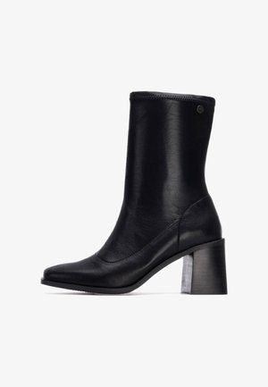 Botín negro de cuero hasta el tobillo con punta afilada, textura suave y tacón grueso. Presenta un diseño elegante y un borde superior reforzado.