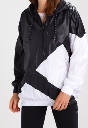 Femme portant un coupe-vent bicolore noir et blanc avec une fermeture éclair à l'avant et une capuche, associé à des leggings noirs.