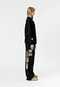 Sudadera negra de gran tamaño con un parche de oso en el pecho, combinada con pantalones negros que cuentan con un parche de oso y un diseño gráfico en el costado.