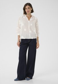 Weiße bestickte Bluse mit floralen Mustern, lockerem Schnitt und langen Ärmeln, kombiniert mit dunklen Jeans mit weitem Bein und blauen Ballerinas.