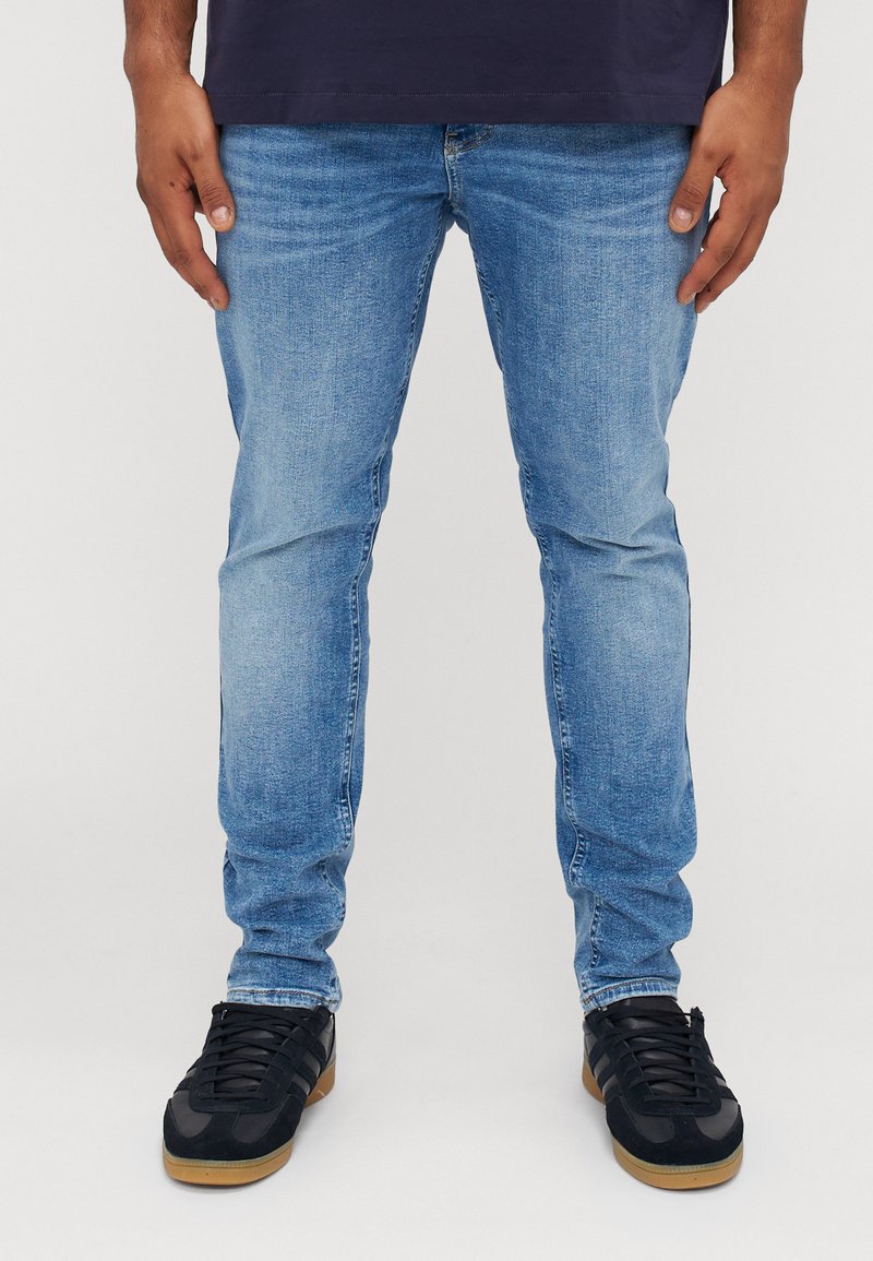 Jeans de mezclilla azul claro con un ajuste slim, que presentan una textura desgastada, diseño de cinco bolsillos y combinados con zapatillas negras sobre un fondo blanco.